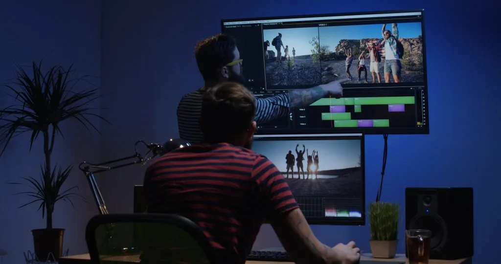 Two individuals are editing video footage on multiple monitors in a dimly lit room