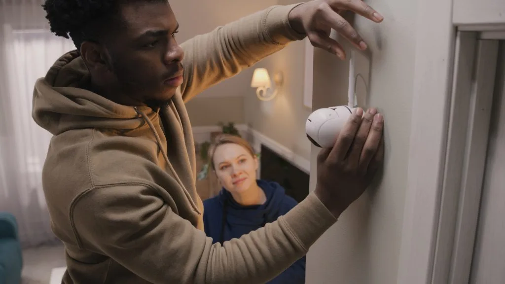 A man installs video security systems on a wall while a woman observes closely