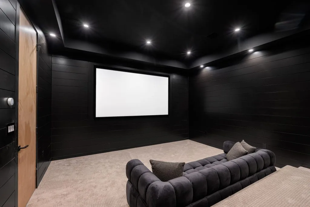 Custom audio visual systems enhance the sleek design of this dark home theater with a large screen and minimalist seating