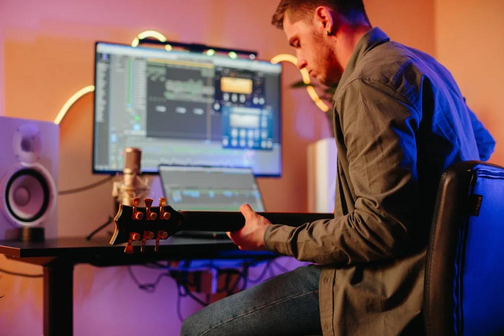 A man plays guitar in a cozy home studio with computer equipment