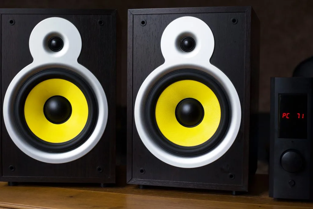 Two speakers with yellow cones sit on a wooden surface next to a control unit