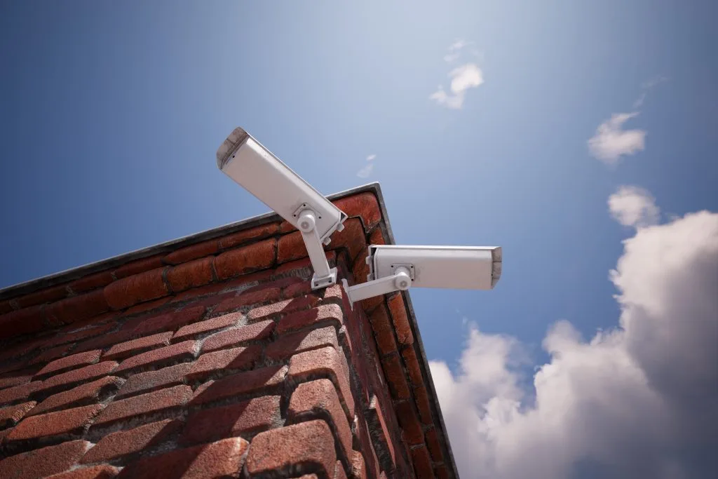 Surveillance cameras are mounted on a brick wall, overlooking a clear sky
