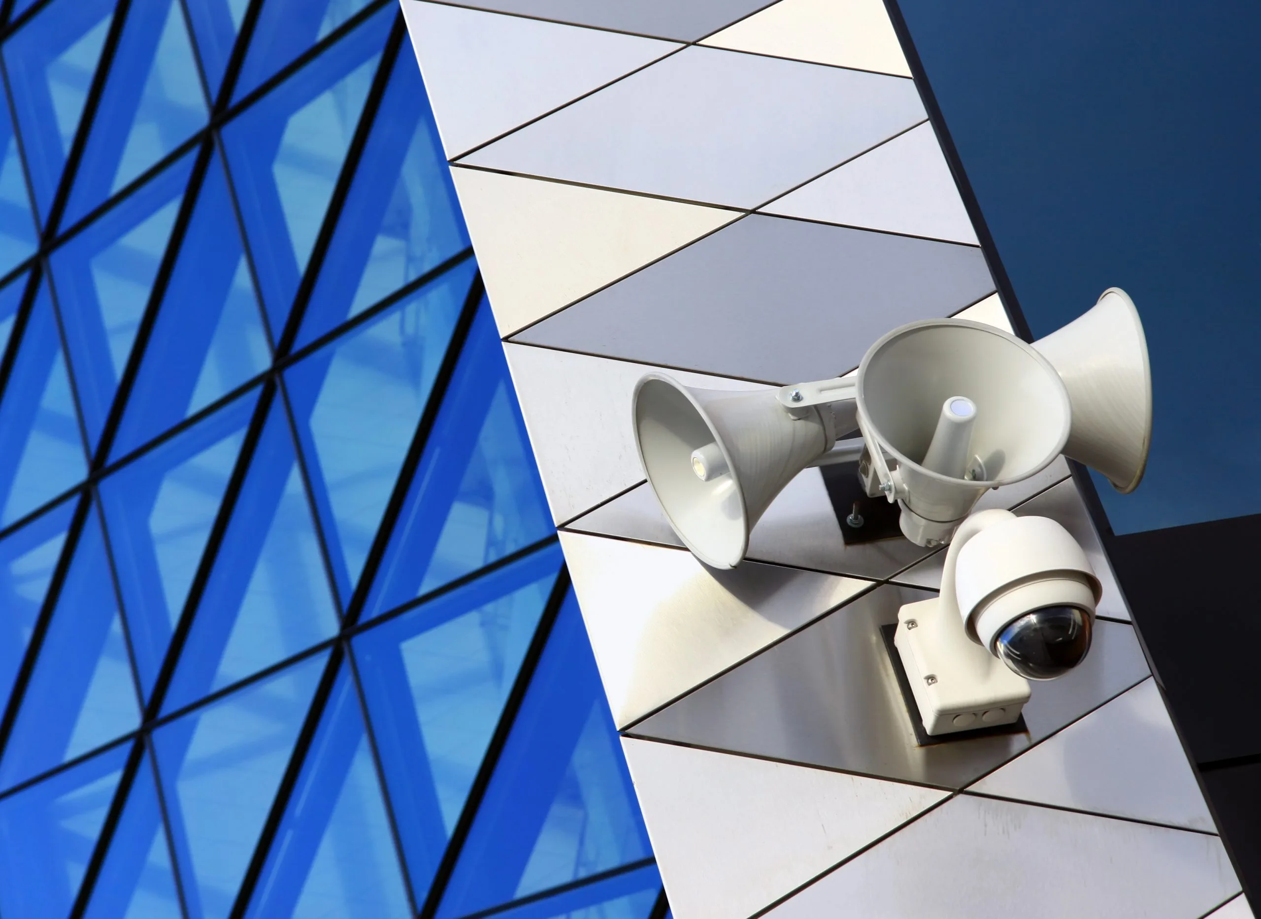 Geometric blue glass reflects in juxtaposition with security cameras and speakers on a wall